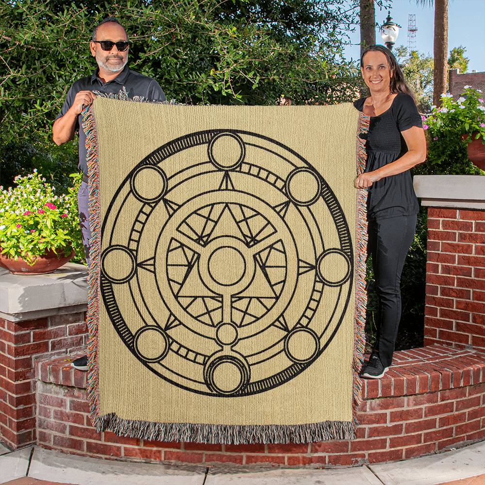 Two people holding up a woven blanket featuring a complex design with these key details: 1. The outermost circle contains eight smaller circles positioned equidistantly along its circumference. These smaller circles are interconnected by straight lines that intersect at the center. 2. Inside this structure, there are additional layers of circles and intricate line patterns, creating a star-like shape in the middle. 3. The entire design is monochromatic, with dark outlines against a lighter background.
